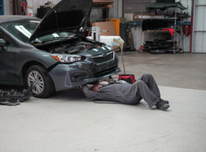 Auto technician lying beneath a raised vehicle performing repair work inside a garage.